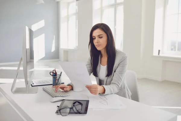 Ernsthaft beschäftigte Geschäftsfrau liest Arbeitsunterlagen am Tisch sitzend mit einem Computer — Stockfoto