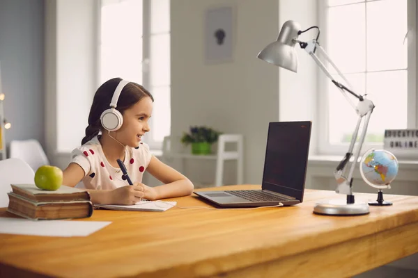 Formación escolar en línea. Chica inteligente en los auriculares está estudiando una lección con un ordenador portátil sentado en una mesa en casa . —  Fotos de Stock