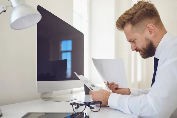 Empresário ocupado sério lê documentos de trabalho sentados em uma mesa com um computador no escritório . — Fotografia de Stock