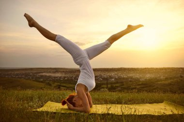 Beyaz spor giysili bir kadın gün batımında çimlerin üzerinde kafasının üstünde yoga balsası yapıyor..