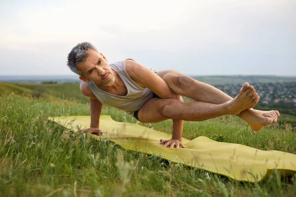 Male yoga on the grass in summer. Mature gray-haired man practicing yoga balance pose in nature