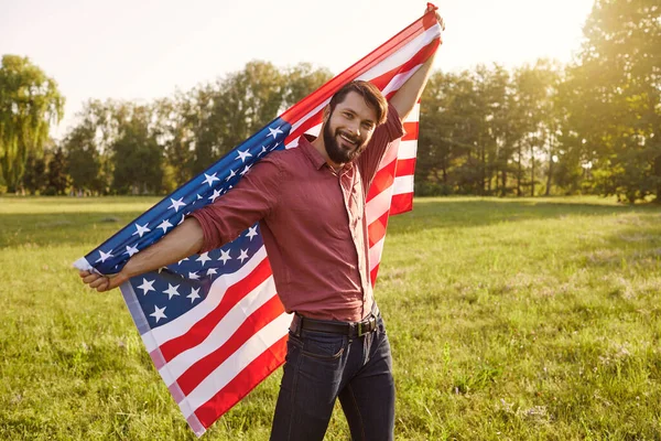 Hombre barbudo hombre con la bandera americana en sus manos al atardecer en el sol . — Foto de Stock