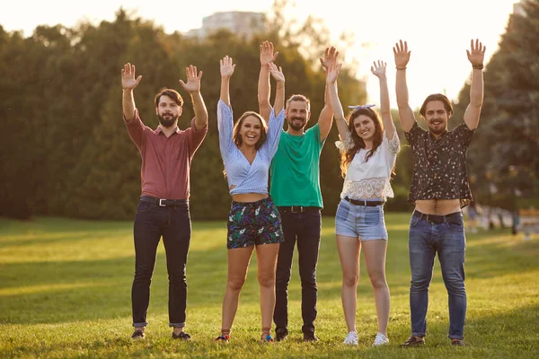 Gelukkige grappige vrienden hieven hun handen op voor een picknick in een zomerpark bij zonsondergang. — Stockfoto