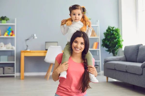 Улыбающаяся мать носит милую маленькую дочку на плечах дома. Родители и дети с игрушками, играющими в помещении — стоковое фото