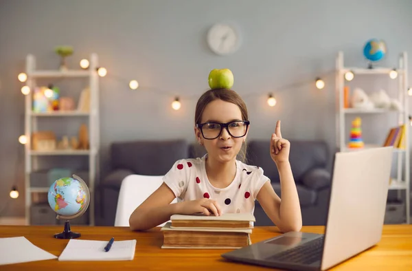 Adorable niña con manzana en la cabeza y un montón de libros apuntando hacia arriba en frente de la computadora portátil en casa — Foto de Stock