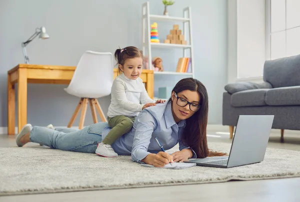 Multitasking för arbete hemma mammor. Ung kvinna gör online-jobb på laptop medan hennes dotter sitter på rygg — Stockfoto