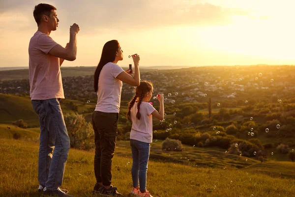 Família afetuosa se divertindo soprando bolhas de sabão no topo da montanha ao pôr do sol. Pais e filhos brincando juntos — Fotografia de Stock