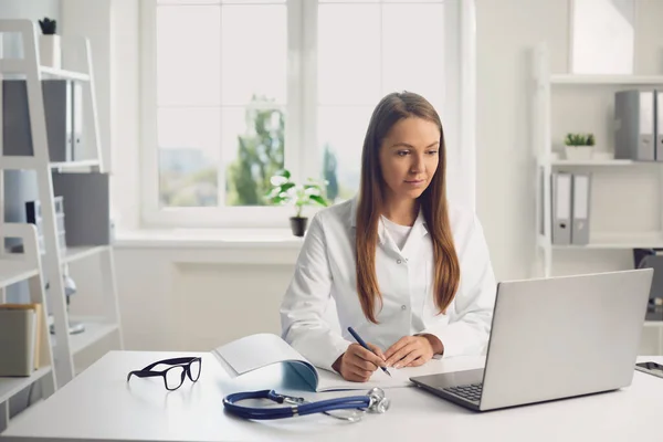 Die Ärztin, die einem Patienten zuhört, sitzt mit einem Laptop an einem Tisch in der Praxis einer Klinik. Online-Konsultation eines Kinderarztes per Laptop-Videokonferenz. — Stockfoto