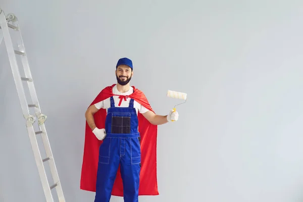 Glad hem reparatör engagerad i hus renovering på grå bakgrund. Ung entreprenör håller roller borste, bär superhjälte kostym nära grå vägg. — Stockfoto