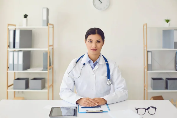 Hispanische Ärztin schaut in die Kamera und hört einer Patientin zu, die eine Webcam-Konferenz nutzt, während sie an einem Tisch im Büro einer medizinischen Klinik sitzt. — Stockfoto