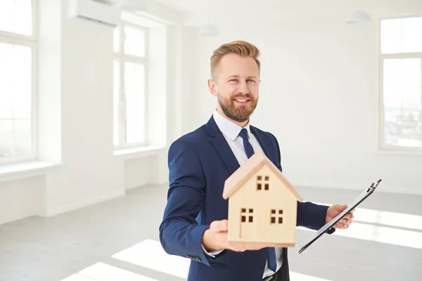 Agente de bienes raíces sosteniendo un modelo de la casa sonriendo en casa habitación blanca de bienes raíces . —  Fotos de Stock