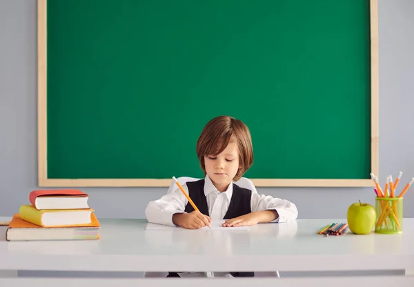 Повернутися до школи. Смішний маленький хлопчик з довгим волоссям пише в блокноті за столом на фоні шкільної зеленої дошки — стокове фото
