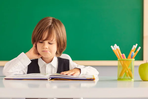 De vuelta a la escuela. Un escolar está leyendo un libro mientras está sentado en la mesa por primera vez en la escuela en el fondo de la pizarra escolar . — Foto de Stock