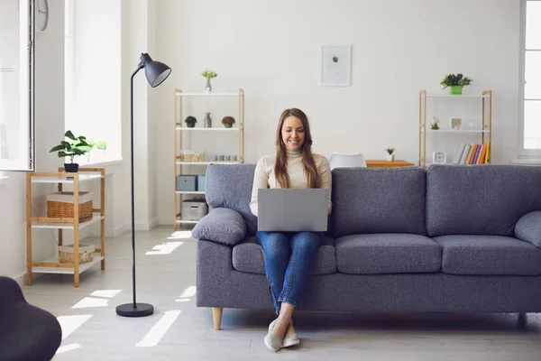 Giovane bella ragazza utilizza un computer portatile mentre si siede su un divano nell'appartamento. Donna lavora ufficio a casa. — Foto Stock