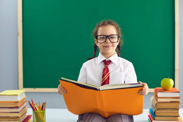 Terug naar school. Slim schoolmeisje in bril met een boek in haar handen zittend op de achtergrond van het schoolbord. — Stockfoto