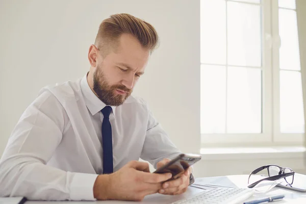 Empresário ocupado sério lê mensagem no telefone em uma mesa com um computador no escritório . — Fotografia de Stock