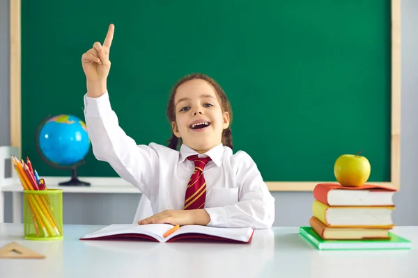 Vissza a suliba. Fogantatás ötlet az iskolában. Vidám iskoláslány felemelte az ujját, miközben egy zöld iskolatáblához ült az osztályteremben.. — Stock Fotó