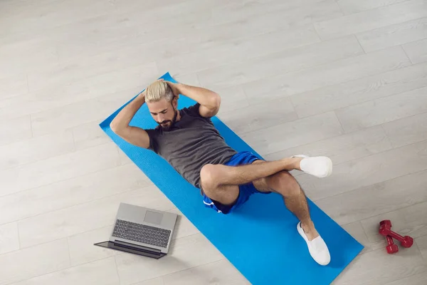 Молодой фитнес-человек делает упражнения на животе, лежа на полу, смотрит онлайн-курс тренера, имея ноутбук в комнате. — стоковое фото