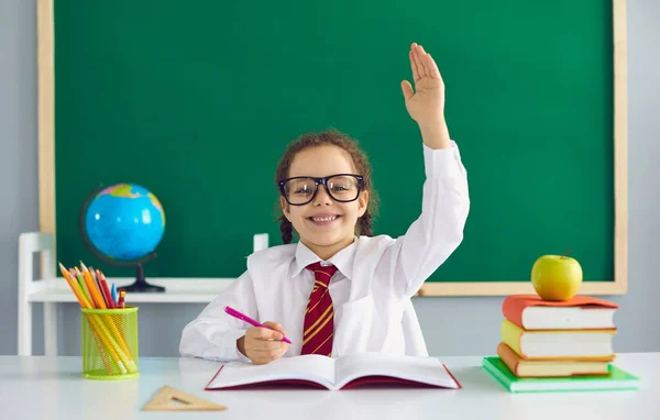 Eerste schooldag. Een gelukkig schoolmeisje met een glimlachende bril stak haar hand op terwijl ze aan een tafel in de klas zat tegen de achtergrond van het schoolbord. — Stockfoto