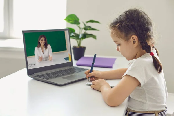 Онлайн школьное образование. Маленькая девочка, которую школьница пишет в блокноте, имеет видео-урок с учительницей ноутбука дома . — стоковое фото