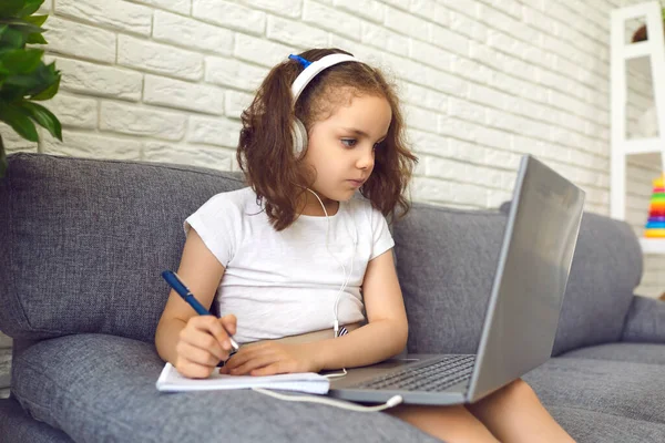 Онлайн-образование ребенка. Очаровательный ребенок в наушниках смотрит видео уроки на ноутбуке и делает заметки дома — стоковое фото