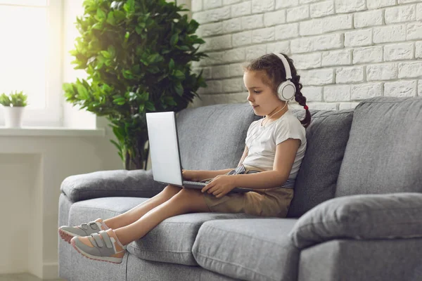 Educación en línea de los niños. Dulce chica con auriculares mirando video lección profesor conferencia portátil sentado en el sofá en casa . —  Fotos de Stock