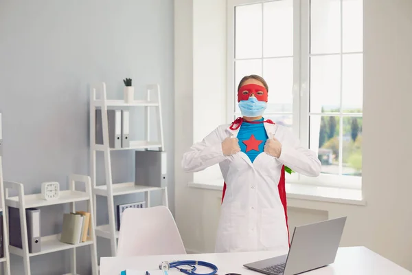 Online medicine. Young female doctor opening her uniform to reveal superhero costume near laptop at her office