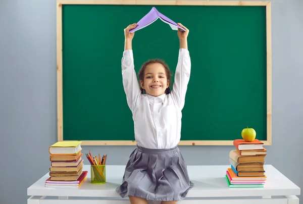 Terug naar school. Vrolijk klein meisje zit op het bureau en houdt boek boven haar hoofd in de klas. Schattig kind hebben plezier op school — Stockfoto