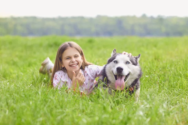Очаровательная маленькая девочка с собакой Хаски, лежащей на зеленом лугу снаружи. Прекрасный ребенок с домашним животным в сельской местности — стоковое фото