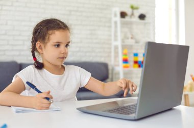 Online eğitim. Çocuklar için video dersi okulu. Küçük bir kız laptopta video dersi izliyor. Odadaki masada oturuyor..