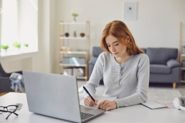 Uzaktan kumanda. Genç bayan serbest çalışan masada oturuyor ve proje üzerinde çalışıyor. Fikirlerini deftere yazıyor.
