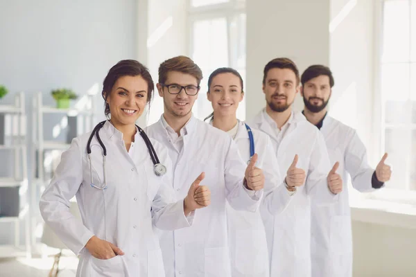 Młodzi uśmiechnięci lekarze zespół portret z kciukami w górę gest w nowoczesnym gabinecie klinicznym. — Zdjęcie stockowe