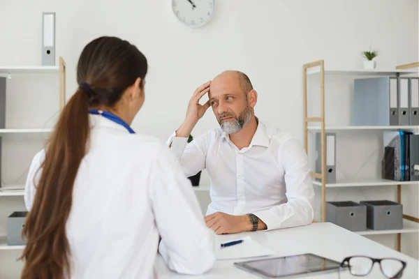 Uomo maturo con mal di testa consulenza con il medico femminile in clinica. Specialista medico che aiuta il paziente a curare l'emicrania — Foto Stock