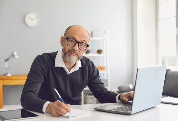 Een oudere man met een bril werkt hij studeert hij heeft een laptop hij schrijft notities in een notitieboekje terwijl hij aan een tafel zit in zijn kantoor. — Stockfoto