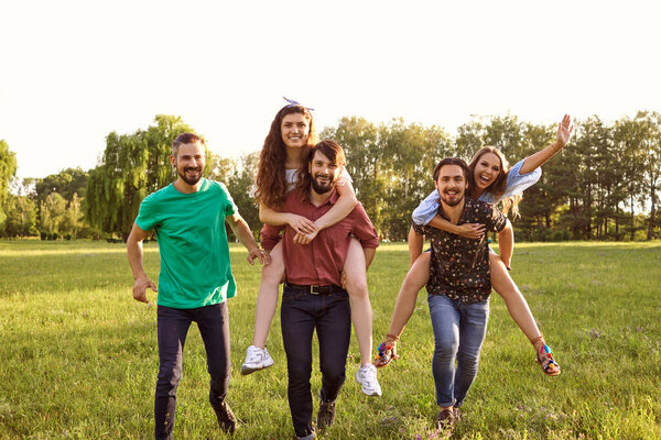 Group of friends walking in countryside with guys giving piggyback ride to girls. Young people having fun in nature