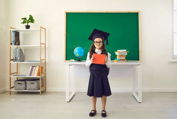 Je retourne à l'école. Une jolie écolière dans un chapeau d'études supérieures avec des livres se dresse dans le contexte d'une classe dans une école. — Photo