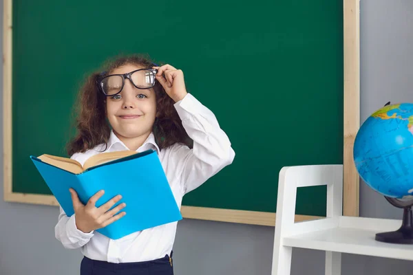 Torna a scuola.Studentessa intelligente in occhiali con un libro in mano sullo sfondo della lavagna della scuola — Foto Stock