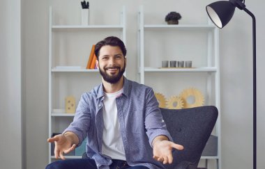 Kameraya bakan beyaz bir adam oturma odasında dostça bir hoş geldin jesti olarak kollarını açıp görüntülü sohbet ediyor..