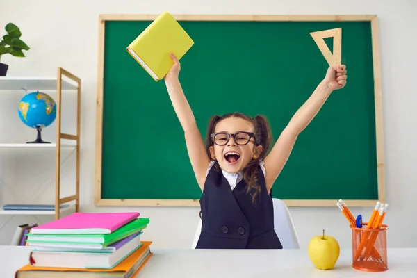Terug naar school. Klein gelukkig schoolkind stak de handen op tegen de achtergrond van een groen schoolbord in een klaslokaal. — Stockfoto