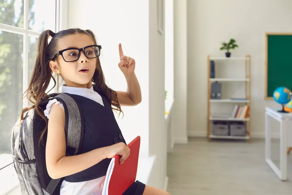 Vissza a suliba. Az iskoláslány szemüvegben, hátizsákkal felemelte az ujját az osztály hátterében ülve.. — Stock Fotó