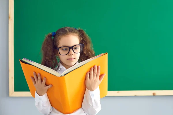 Back to school. Smart little girl in glasses reading huge book near blackboard at classroom. Cute first grader studying at school