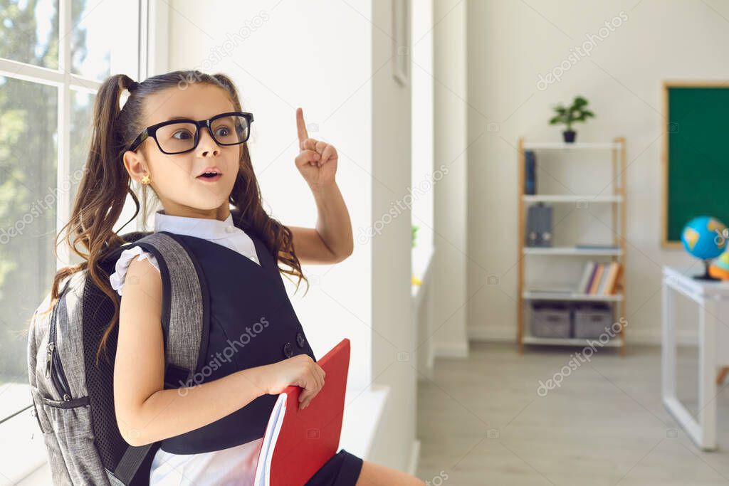 Back to school. The schoolgirl in glasses with a backpack raised her finger up sitting on a background of class.