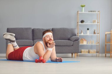 Funny red fat man doing exercises on the floor smiling while lying on the floor at home.