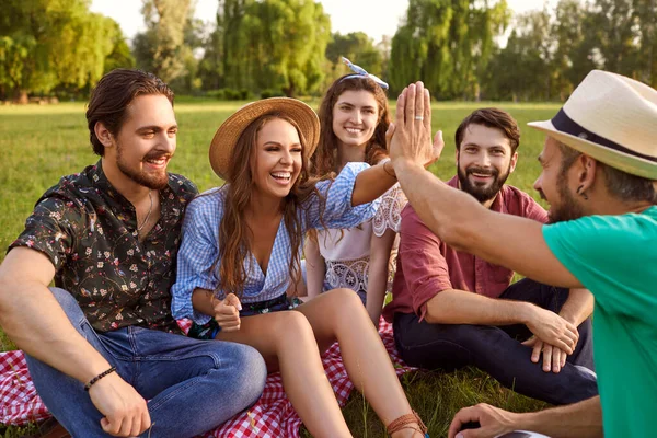 Group of friends sitting on grass, talking and high fiving in countryside. Joyful people enjoying time together outdoor in park — Stock Photo, Image