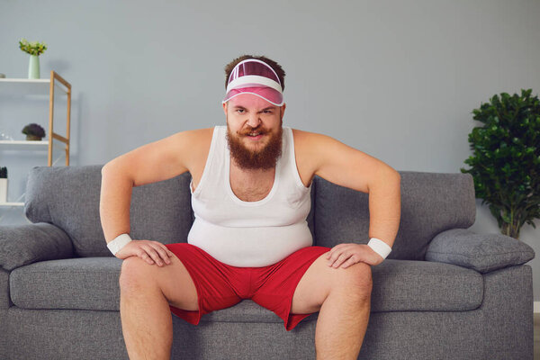 Funny fat man in sportswear is sitting on the sofa in the room