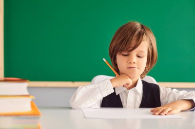 Back to school. Serious little boy writing something in notebook at desk in classroom, copy space. Hardworking child studying at school clipart