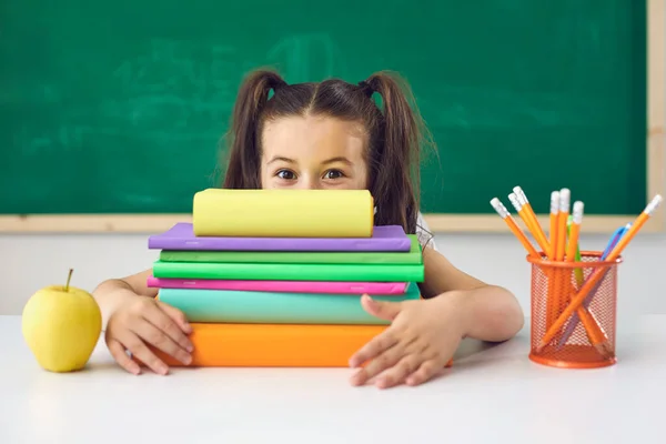 Grappig klein schoolmeisje houdt een boek boven haar hoofd tijdens het zitten in een les in de klas op school. — Stockfoto