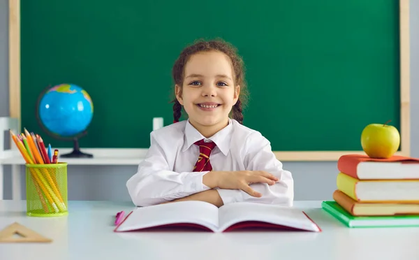 Концепція повернення до школи. Щаслива позитивна школярка посміхається, сидячи за столом на фоні шкільної дошки в класі . — стокове фото