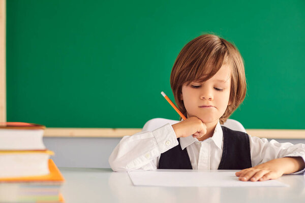 Back to school. Serious little boy writing something in notebook at desk in classroom, copy space. Hardworking child studying at school