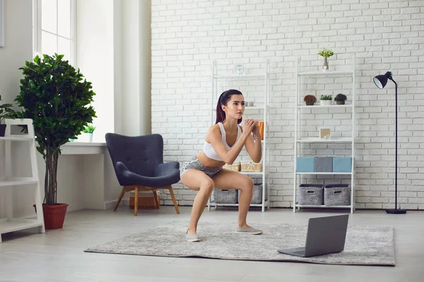 Mujer joven fuerte haciendo ejercicio a video tutorial en línea en casa. Deportiva chica haciendo sentadillas durante su entrenamiento en interiores — Foto de Stock